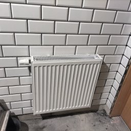 MIM INSTALACJE - Biały grzejnik panelowy zamontowany na ścianie wyłożonej białymi, prostokątnymi płytkami typu metro. Widoczny zawór termostatyczny i fragment drabiny malarskiej.