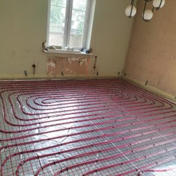 MIM INSTALACJE - Równomiernie ułożone, czerwone rury ogrzewania podłogowego na folii izolacyjnej w jasnym pomieszczeniu z oknem i lampą sufitową. Widoczne ślady po pracach wykończeniowych.