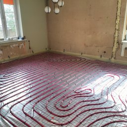 MIM INSTALACJE - Czerwone rury ogrzewania podłogowego ułożone na srebrnej folii w pokoju z oknami i lampą wiszącą. Widoczne ślady po remoncie na ścianach.