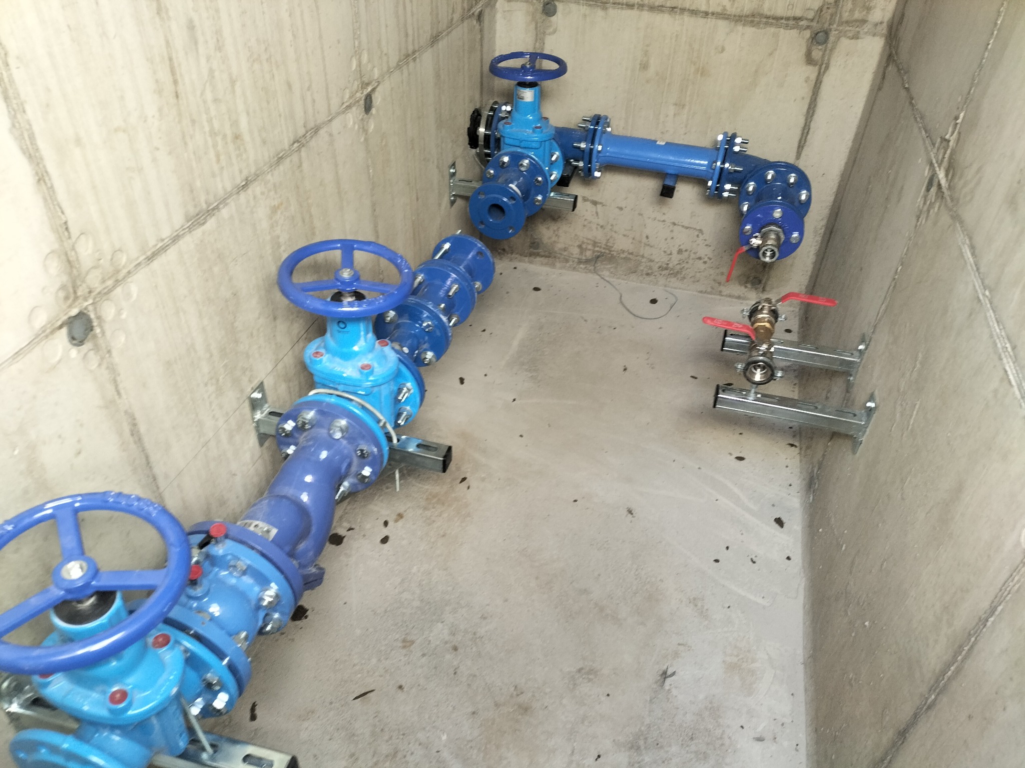 Niebieskie rury z zaworami zamontowane w betonowym pomieszczeniu. Instalacja wodna z metalowymi wspornikami. Widok z góry na gotową instalację.