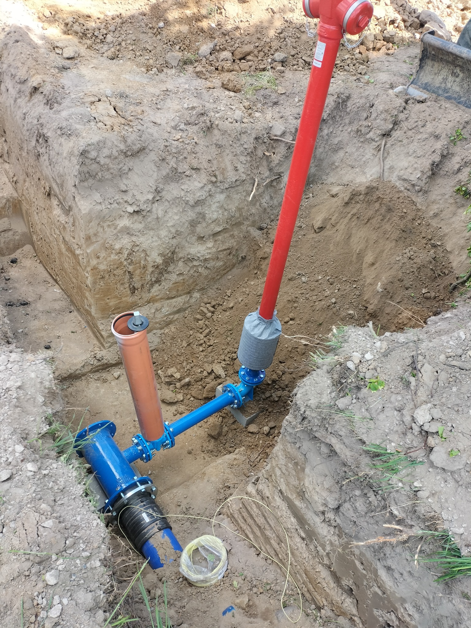 Wykonane przyłącze wod-kan w wykopie ziemnym. Widoczne rury w kolorze niebieskim, czerwonym i pomarańczowym. Elementy złączone, gotowe do zasypania.