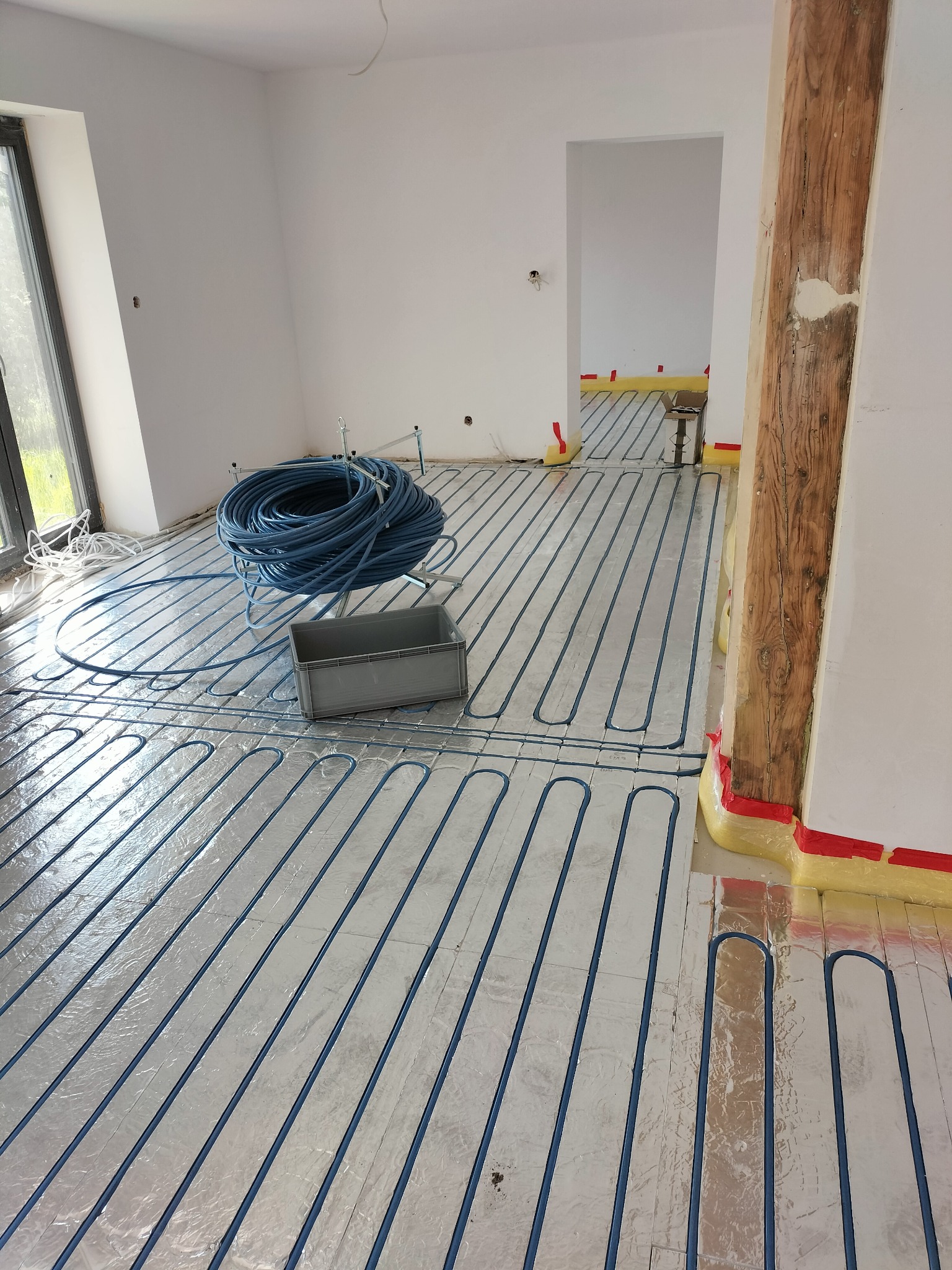 Niebieskie rury ogrzewania podłogowego ułożone na srebrnej folii izolacyjnej w jasnym pomieszczeniu z drewnianym słupem. Część rur zwinięta w krąg obok szarego pojemnika.