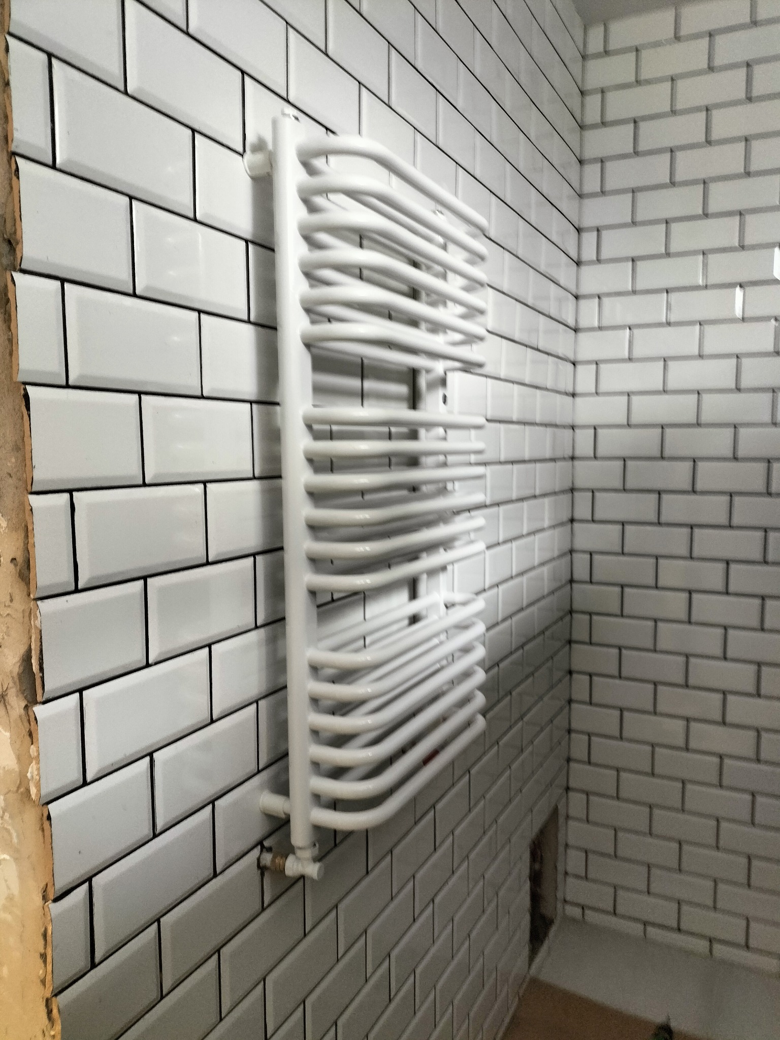 Biały, drabinkowy grzejnik łazienkowy na ścianie z białych, prostokątnych płytek. Widoczne przyłącze. Fragment ściany bez płytek w trakcie prac wykończeniowych.