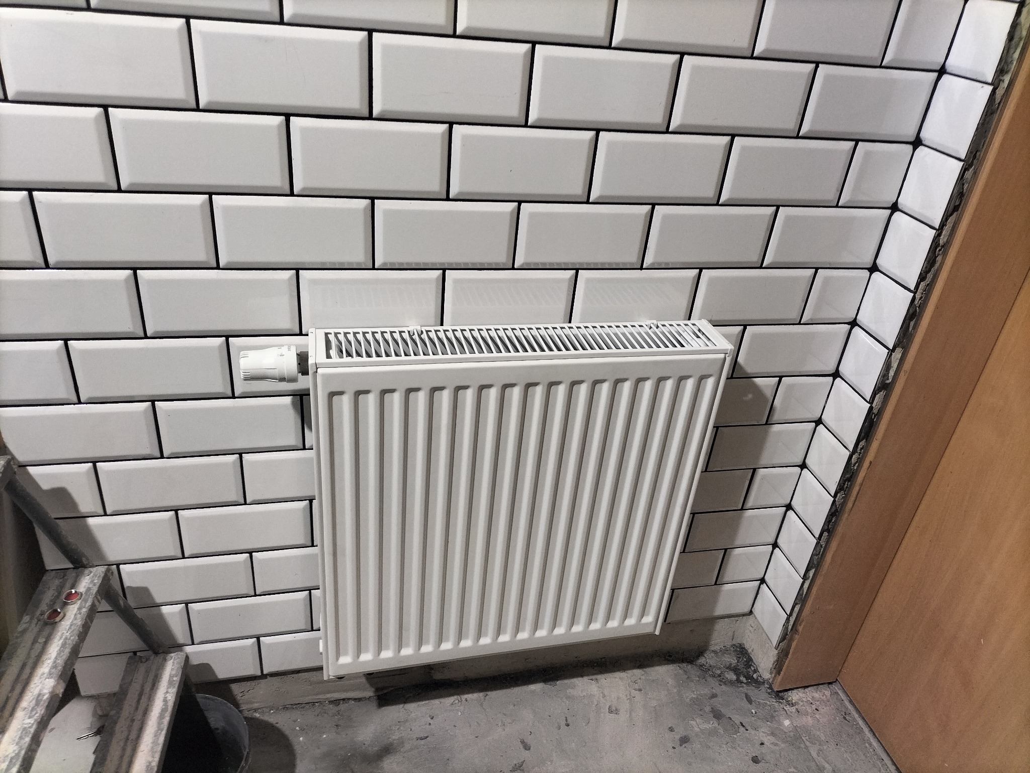Biały grzejnik panelowy zamontowany na ścianie wyłożonej białymi, prostokątnymi płytkami typu metro. Widoczny zawór termostatyczny i fragment drabiny malarskiej.