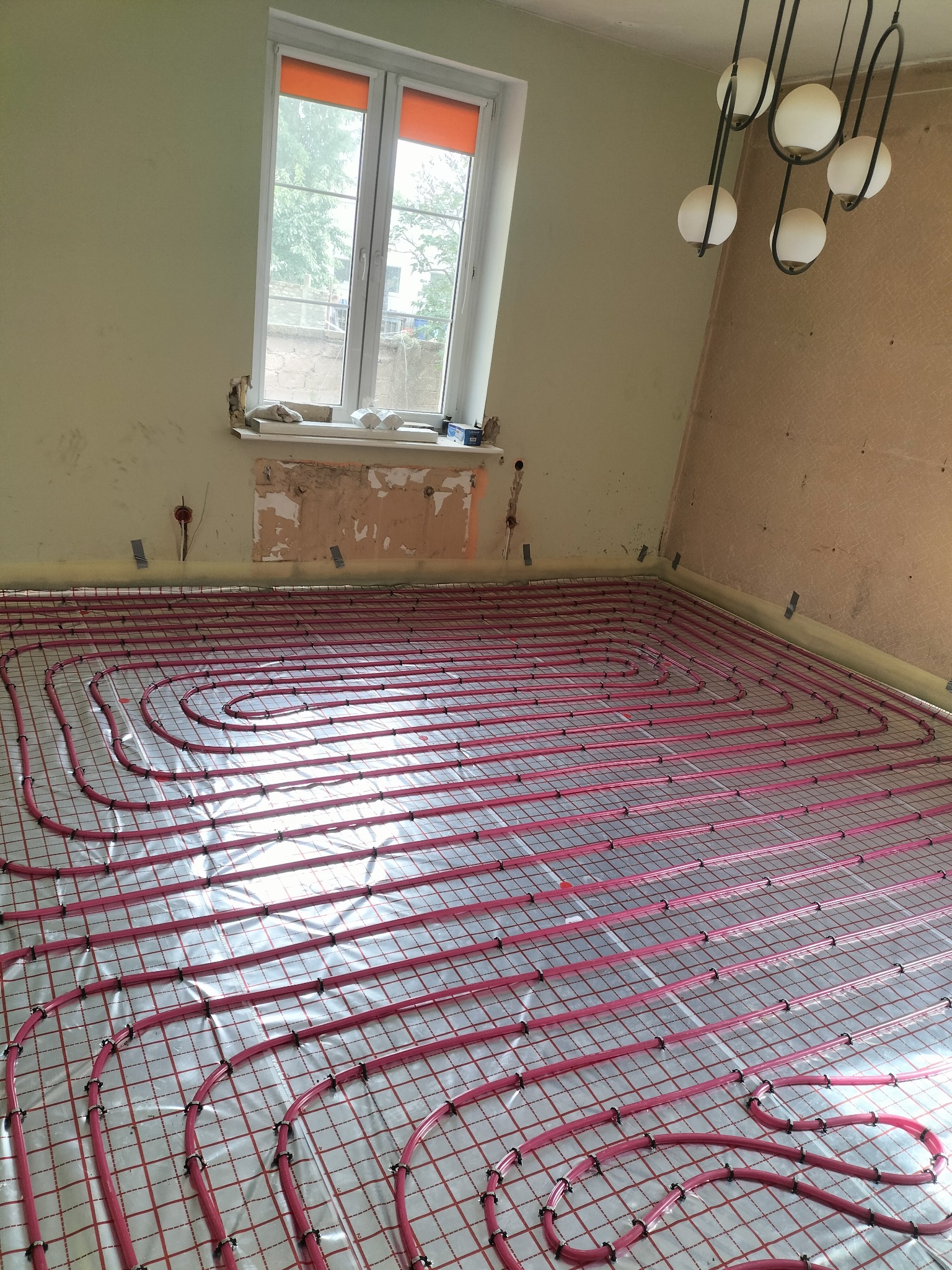 Równomiernie ułożone, czerwone rury ogrzewania podłogowego na folii izolacyjnej w jasnym pomieszczeniu z oknem i lampą sufitową. Widoczne ślady po pracach wykończeniowych.