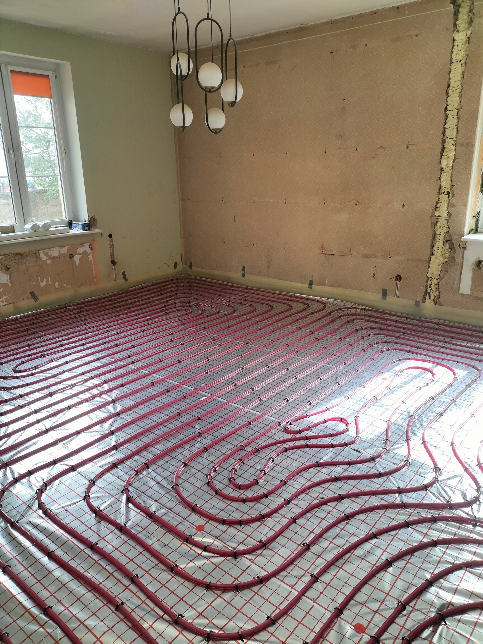 Czerwone rury ogrzewania podłogowego ułożone na srebrnej folii w pokoju z oknami i lampą wiszącą. Widoczne ślady po remoncie na ścianach.