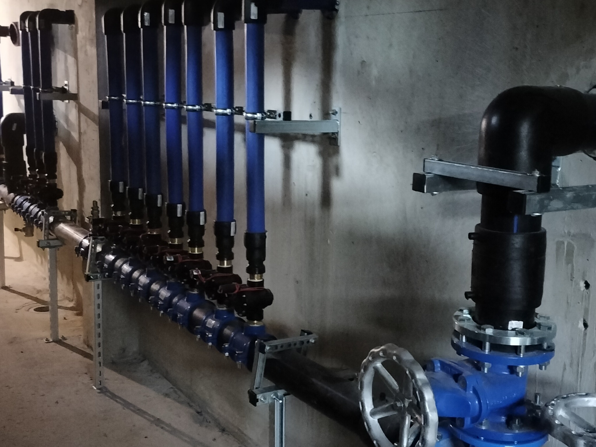 Szereg niebieskich rur instalacji wodnej z zaworami, zamontowanych na metalowych wspornikach przy szarej, betonowej ścianie. Widoczne liczne złącza i elementy instalacyjne.