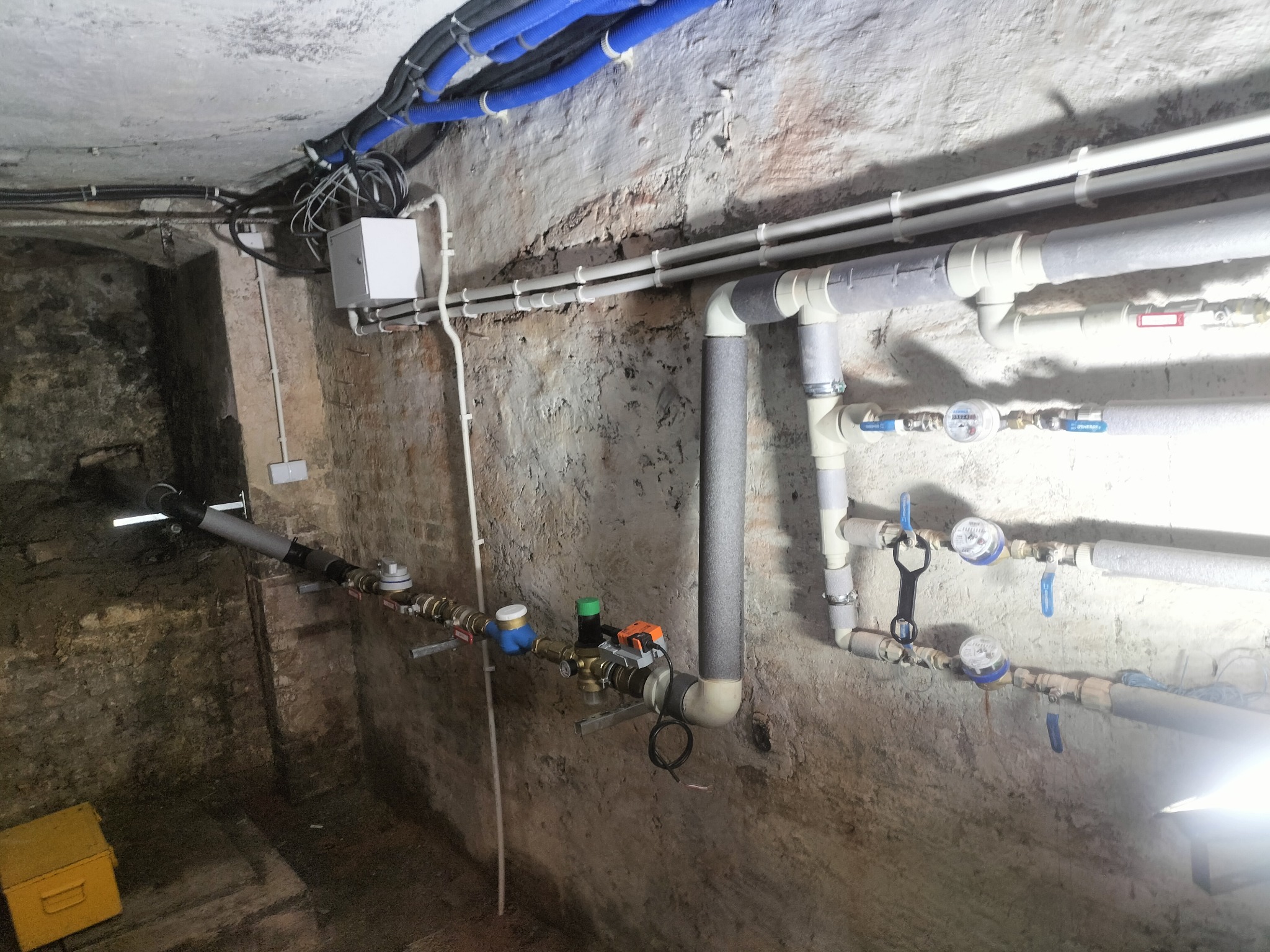 Instalacja wodna z rurami, zaworami i licznikami w piwnicy o surowych ścianach. Widoczne izolacje termiczne na rurach i skrzynka elektryczna na ścianie.