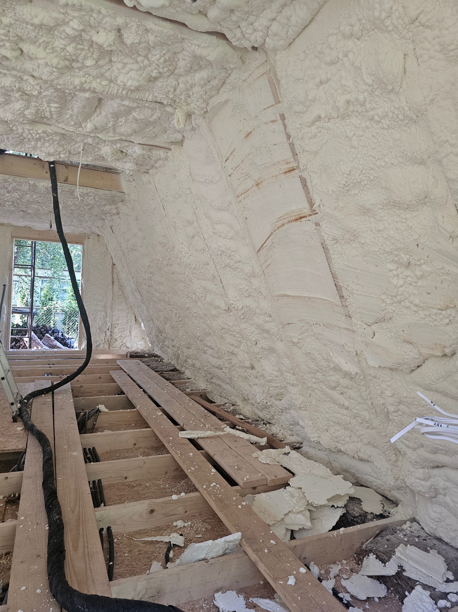 Poddasze z drewnianą konstrukcją i oknem, pokryte grubą warstwą białej pianki izolacyjnej, z widocznym przewodem instalacyjnym i fragmentami odpadów pianki.