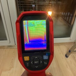 LIVANS SPÓŁKA Z OGRANICZONĄ ODPOWIEDZIALNOŚCIĄ - Pomiar termowizyjny okna termometrem w kolorze czerwonym, wskazujący ucieczkę ciepła. Widoczny fragment okna i podłogi.