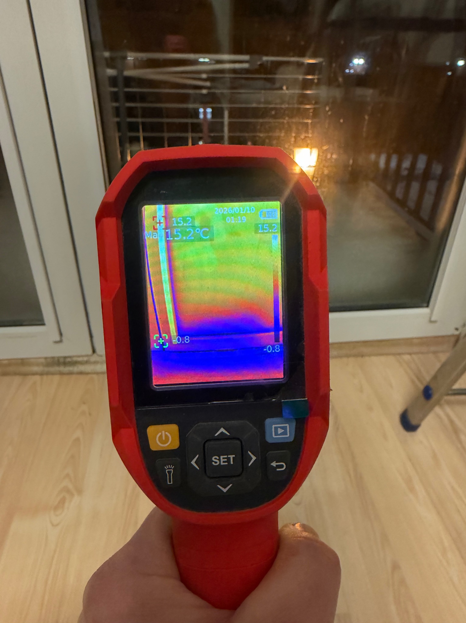 Pomiar termowizyjny okna termometrem w kolorze czerwonym, wskazujący ucieczkę ciepła. Widoczny fragment okna i podłogi.
