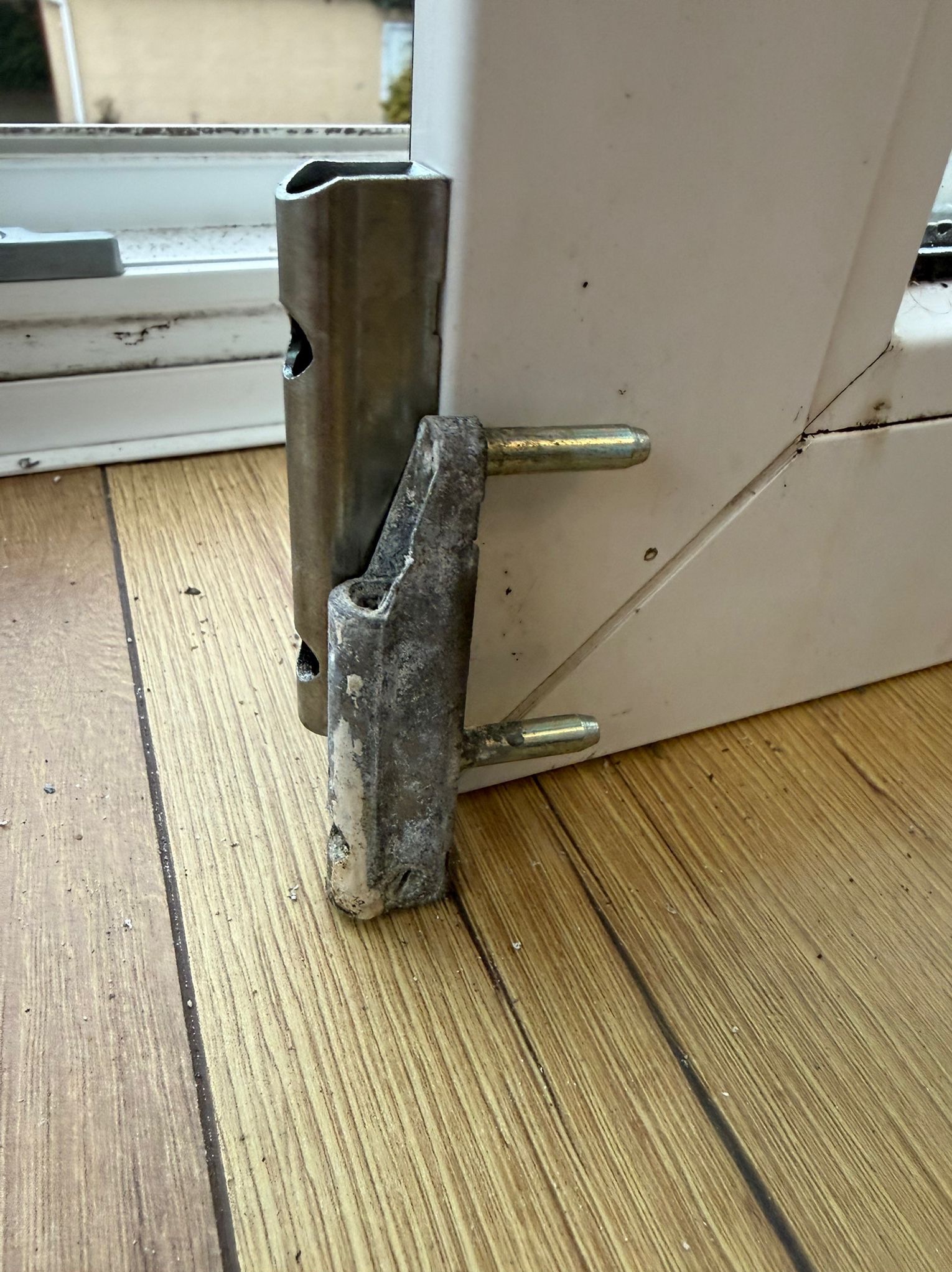 Zardzewiały, skorodowany zawias okienny z widocznymi śladami zużycia, przymocowany do białej ramy okna na tle drewnianej podłogi. Detal uszkodzonego okucia.
