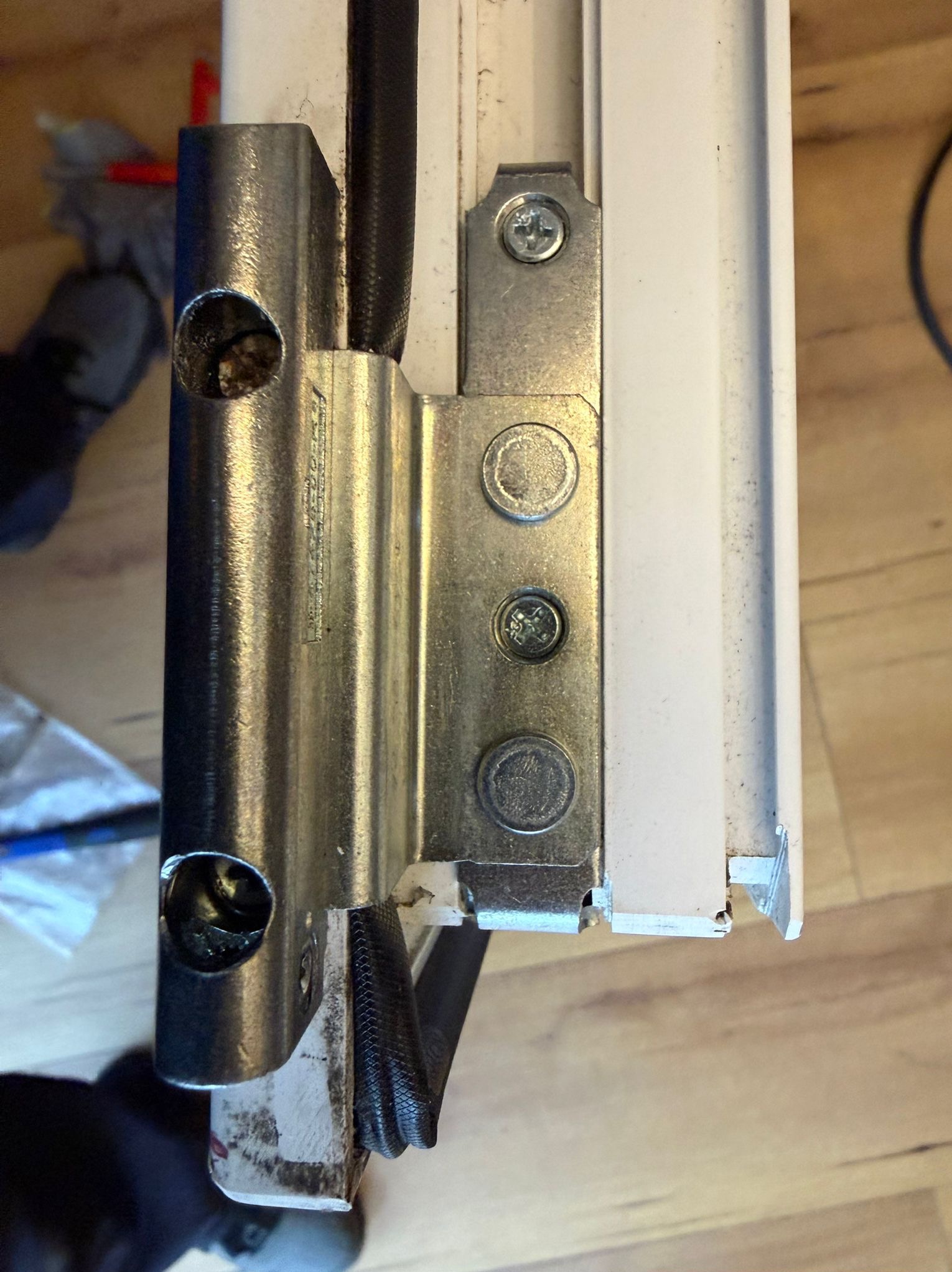 Szczegółowe zbliżenie na zawias okienny przymocowany do białej ramy okna, widoczne śruby mocujące i elementy mechanizmu. Prezentacja różnych typów zawiasów okiennych.