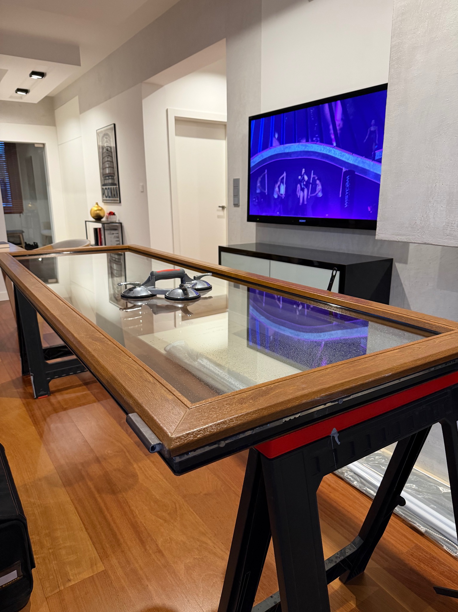 Okno z szybą, leżące na kozłach, przygotowane do montażu. Widoczne przyssawki do szyb. W tle telewizor i elementy wyposażenia wnętrza.