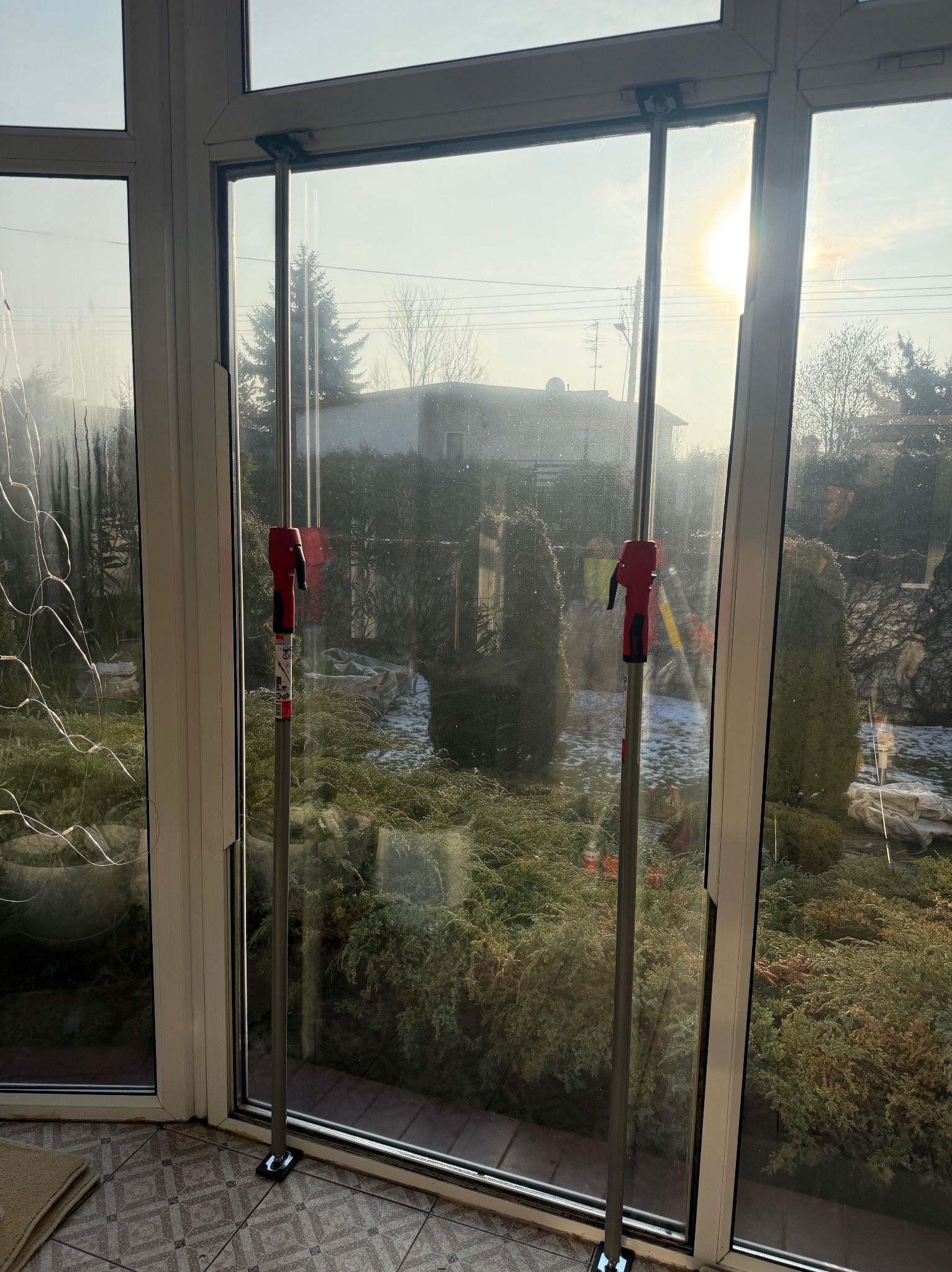 Okno z podporami teleskopowymi w trakcie naprawy, widok na zewnątrz z ogrodem. Słoneczny dzień, częściowo widoczne narzędzia.