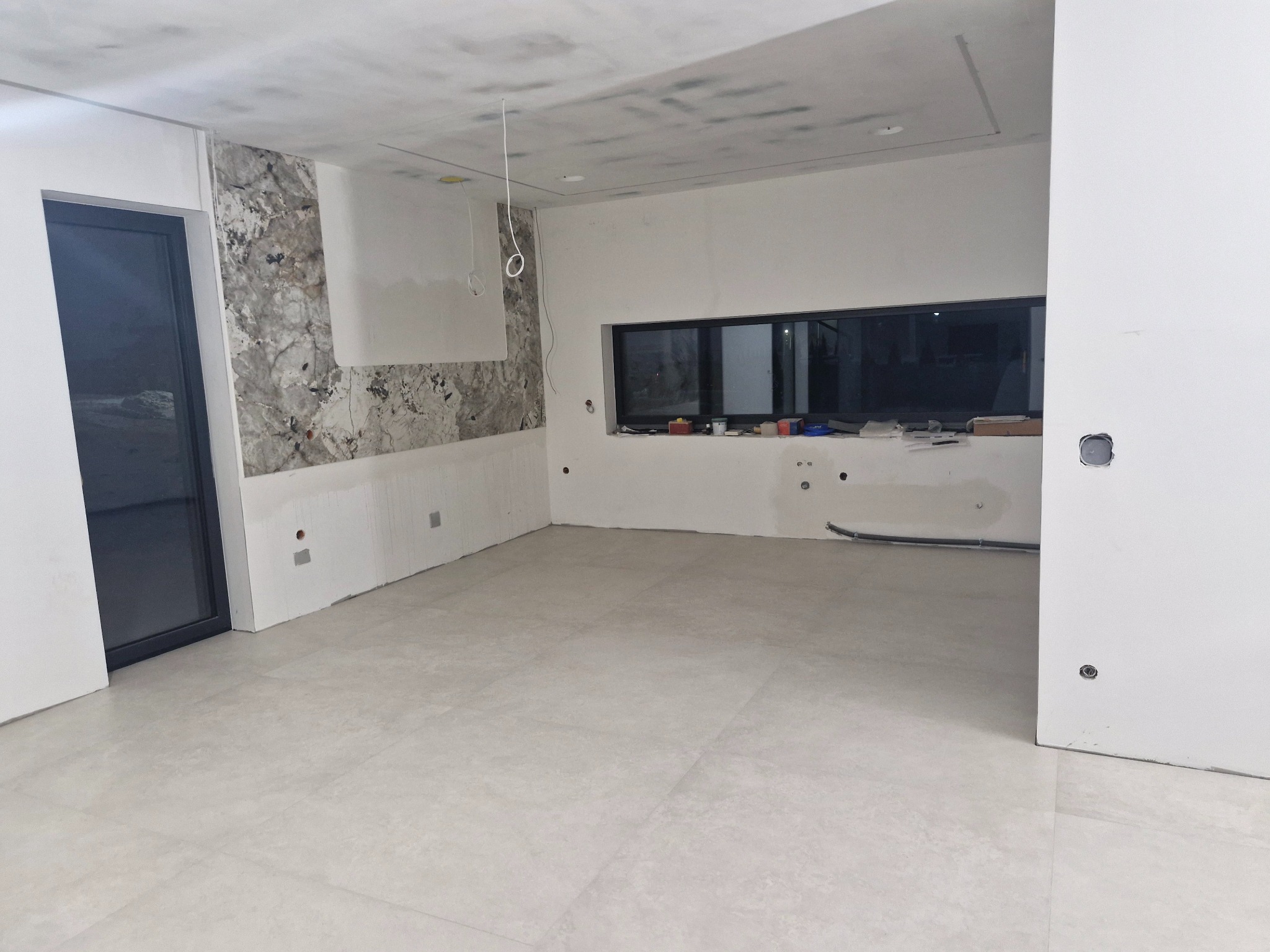 Wnętrze w trakcie wykańczania: położona jasna podłoga, fragment ściany z dekoracyjną okładziną, surowy sufit, okno. Widoczne przewody i puszki elektryczne.