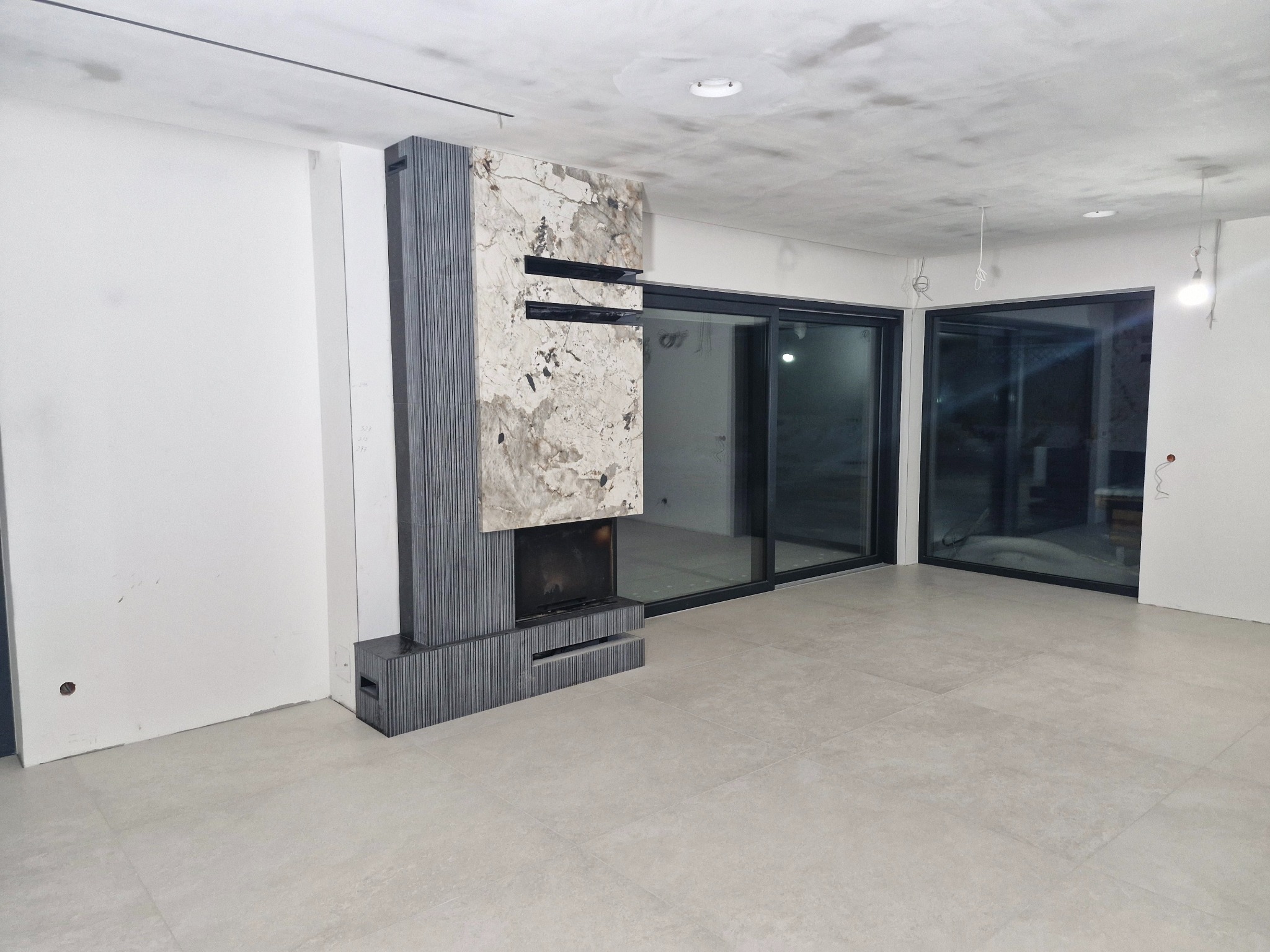 Nowoczesny salon w trakcie wykańczania: jasne płytki podłogowe, kominek obłożony kamieniem i drewnem, duże okna przesuwne. Widoczne niedokończone ściany i sufit.