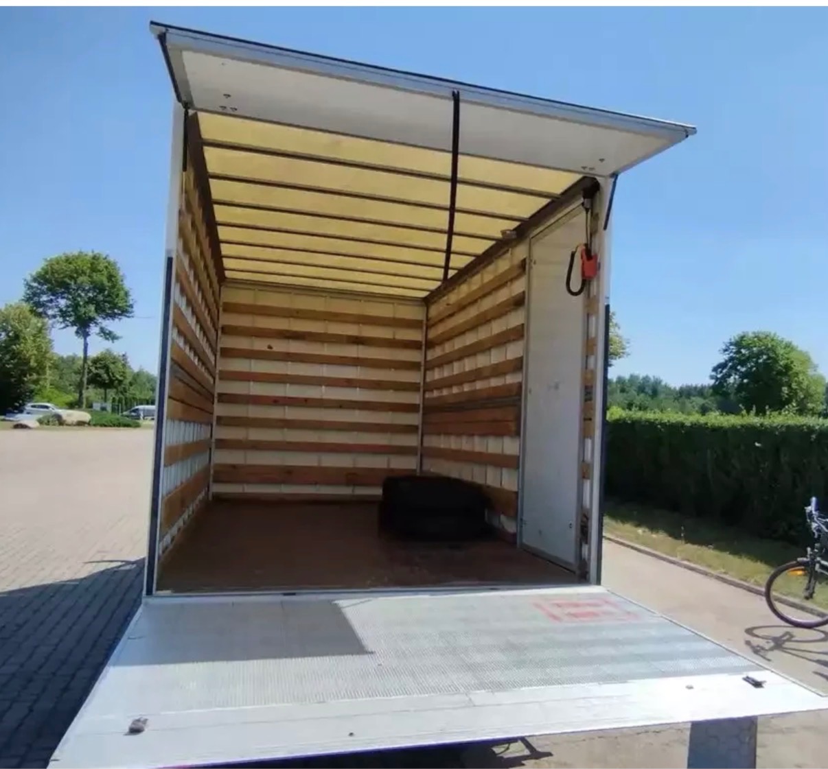 Pusta przestrzeń ładunkowa busa z otwartą klapą, widok z tyłu. Ściany wyłożone drewnem, podłoga aluminiowa. Na podłodze leży opona. Na zewnątrz zieleń i błękitne niebo.