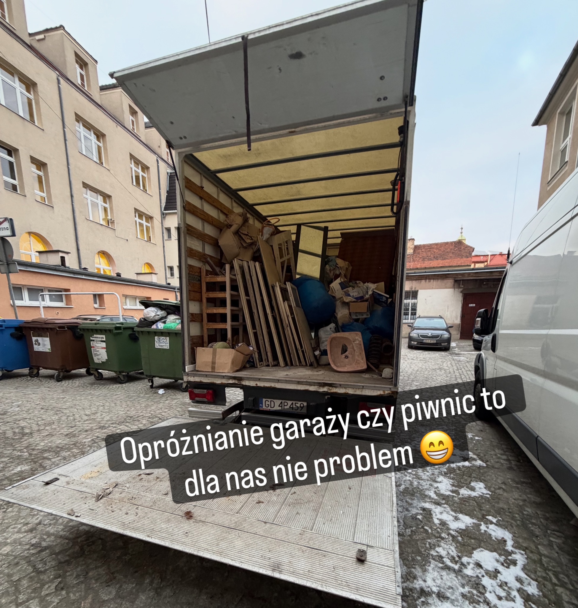 Otwarty bus transportowy wypełniony meblami i przedmiotami z opróżniania garażu/piwnicy, zaparkowany na brukowanym dziedzińcu, z widocznymi kontenerami na śmieci.