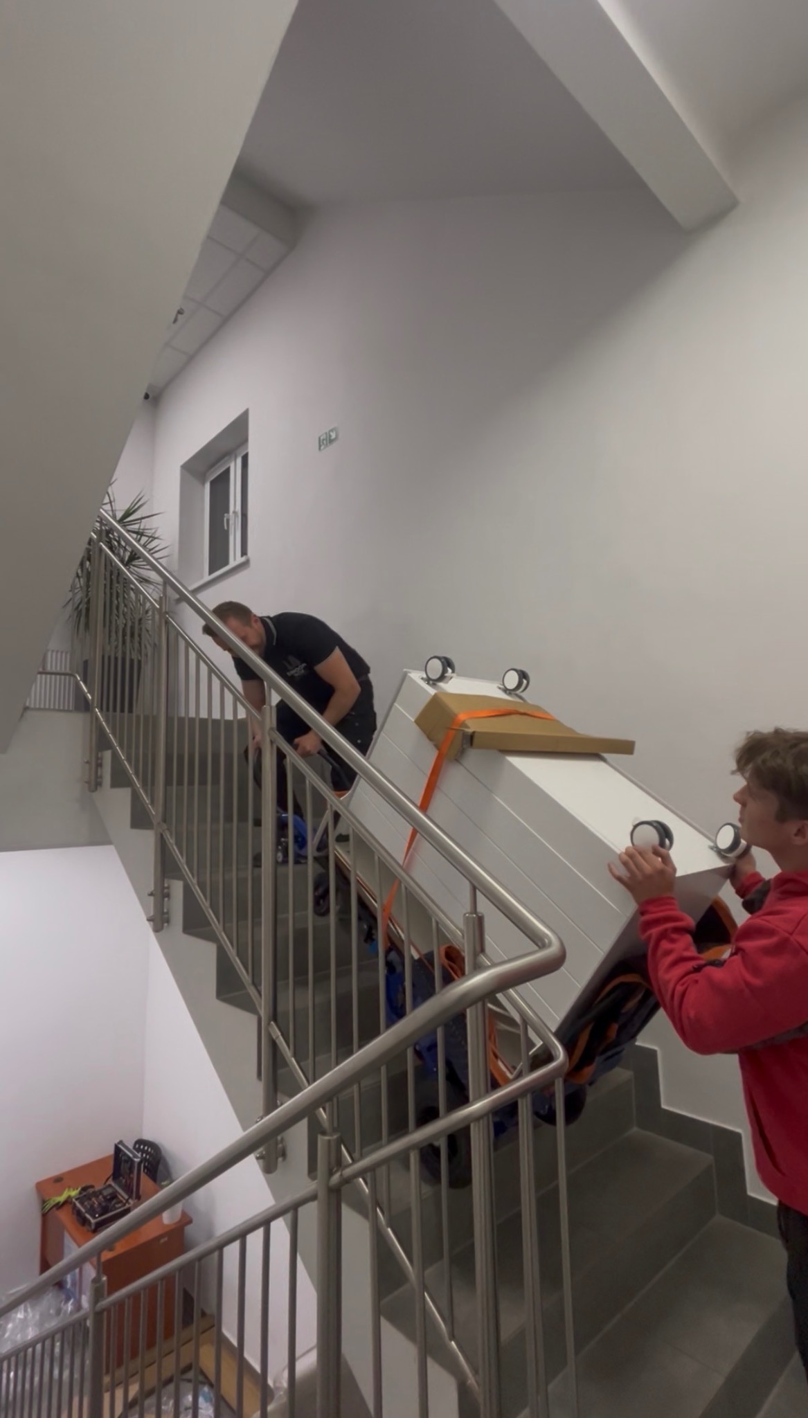 Dwóch mężczyzn wnosi po schodach białą szafę zabezpieczoną pasami transportowymi. Widoczne barierki schodowe i fragment biura z oknem w tle.