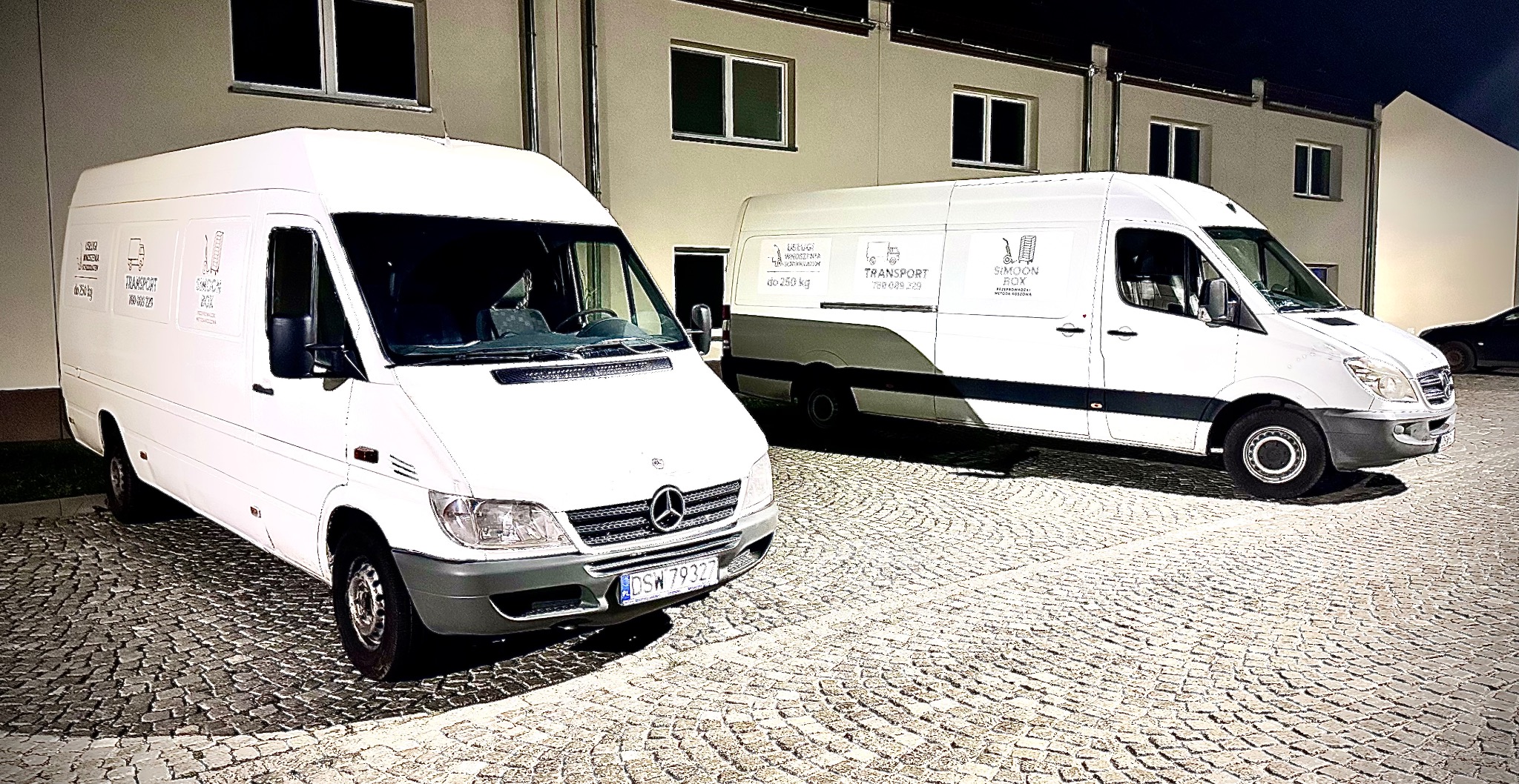 Dwa białe busy transportowe marki Mercedes zaparkowane na brukowanej nawierzchni przed budynkiem. Na busach widoczne logo firmy transportowej.