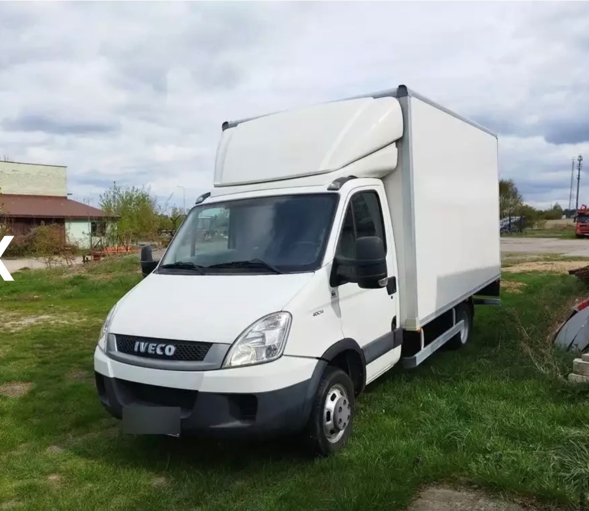 Biały samochód dostawczy Iveco z zabudową kontenerową, zaparkowany na trawie, widok z przodu, pochmurne niebo w tle. Idealny do przeprowadzek i transportu.