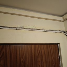 Wikadem Usługi Elektryczne Marcin Jarzyński - Biała rurka instalacyjna przymocowana do ściany nad futryną drzwi, widoczne ślady po kuciu w ścianie. Etap prac instalacyjnych wewnątrz budynku.