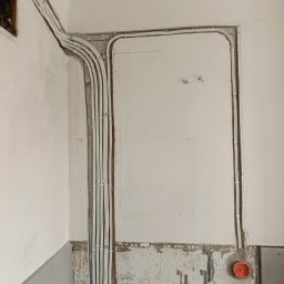 Wikadem Usługi Elektryczne Marcin Jarzyński - Białe kable elektryczne ułożone w bruzdach na ścianie w trakcie remontu. Widoczne puszki instalacyjne i fragmenty starej glazury. Prace instalacyjne w trakcie realizacji.