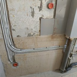 Wikadem Usługi Elektryczne Marcin Jarzyński - Białe kable elektryczne ułożone wzdłuż ściany w trakcie remontu, widoczne puszki instalacyjne i fragment instalacji kanalizacyjnej. Surowy stan wykończenia.