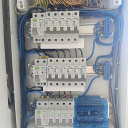 Wikadem Usługi Elektryczne Marcin Jarzyński - Rozdzielnica elektryczna z bezpiecznikami i okablowaniem wewnątrz białej obudowy. Widoczne oznaczenia na przewodach i elementach. Ujęcie z góry, ostre detale.