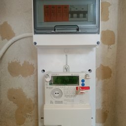 Wikadem Usługi Elektryczne Marcin Jarzyński - Zamontowana skrzynka elektryczna z licznikiem i zabezpieczeniami na ścianie o nierównej fakturze. Widoczne przewody i elementy instalacji.