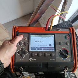Wikadem Usługi Elektryczne Marcin Jarzyński - Miernik Sonel MPI-520 w trakcie pomiaru rezystancji izolacji, z widocznymi przewodami pomiarowymi i wskazaniem wartości na wyświetlaczu. Ustawiony na drabinie.