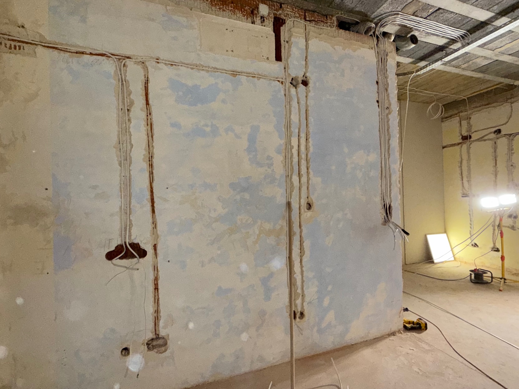 Surowe wnętrze w trakcie prac elektrycznych: kable w bruzdach na ścianie, puszki pod gniazdka, lampa robocza. Widoczne etapy instalacji elektrycznej w budynku.