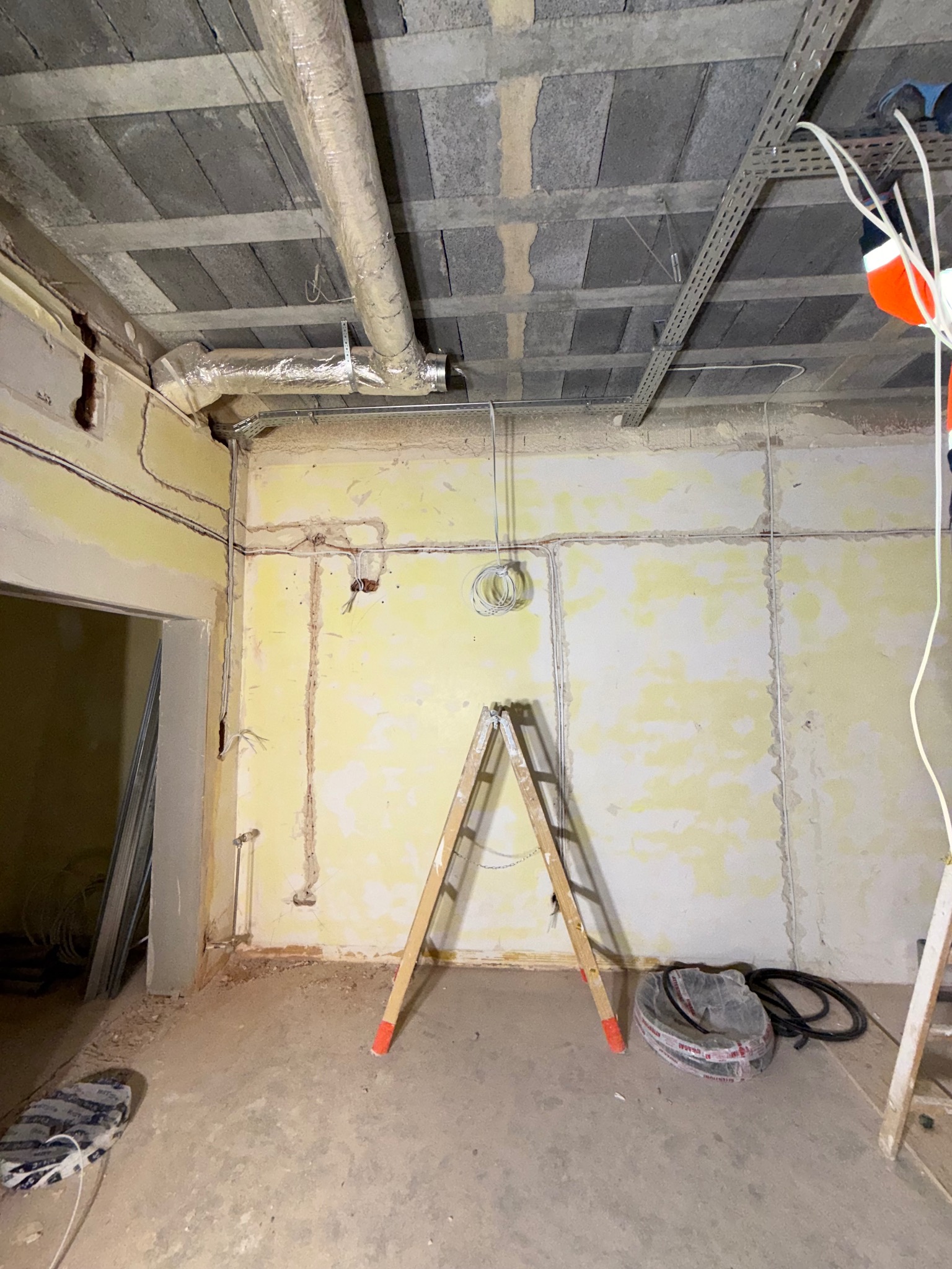Wnętrze w trakcie remontu z widocznymi przewodami elektrycznymi na ścianach, drabina i rolka kabla na podłodze, oraz instalacja wentylacyjna pod betonowym sufitem.