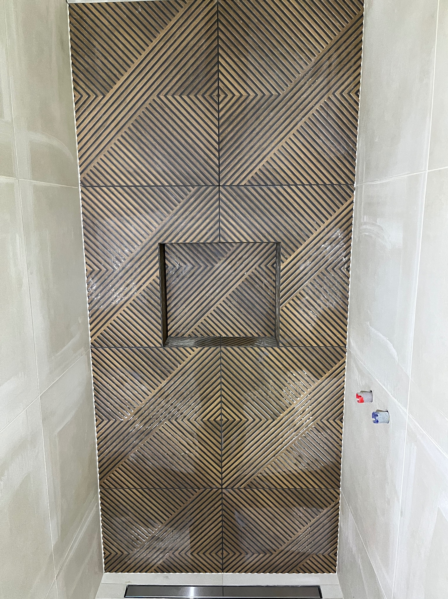 Ściana prysznicowa z brązowo-beżowych płytek ułożonych we wzór geometryczny, z wnęką na kosmetyki i odpływem liniowym. Widoczne przyłącza wodne.
