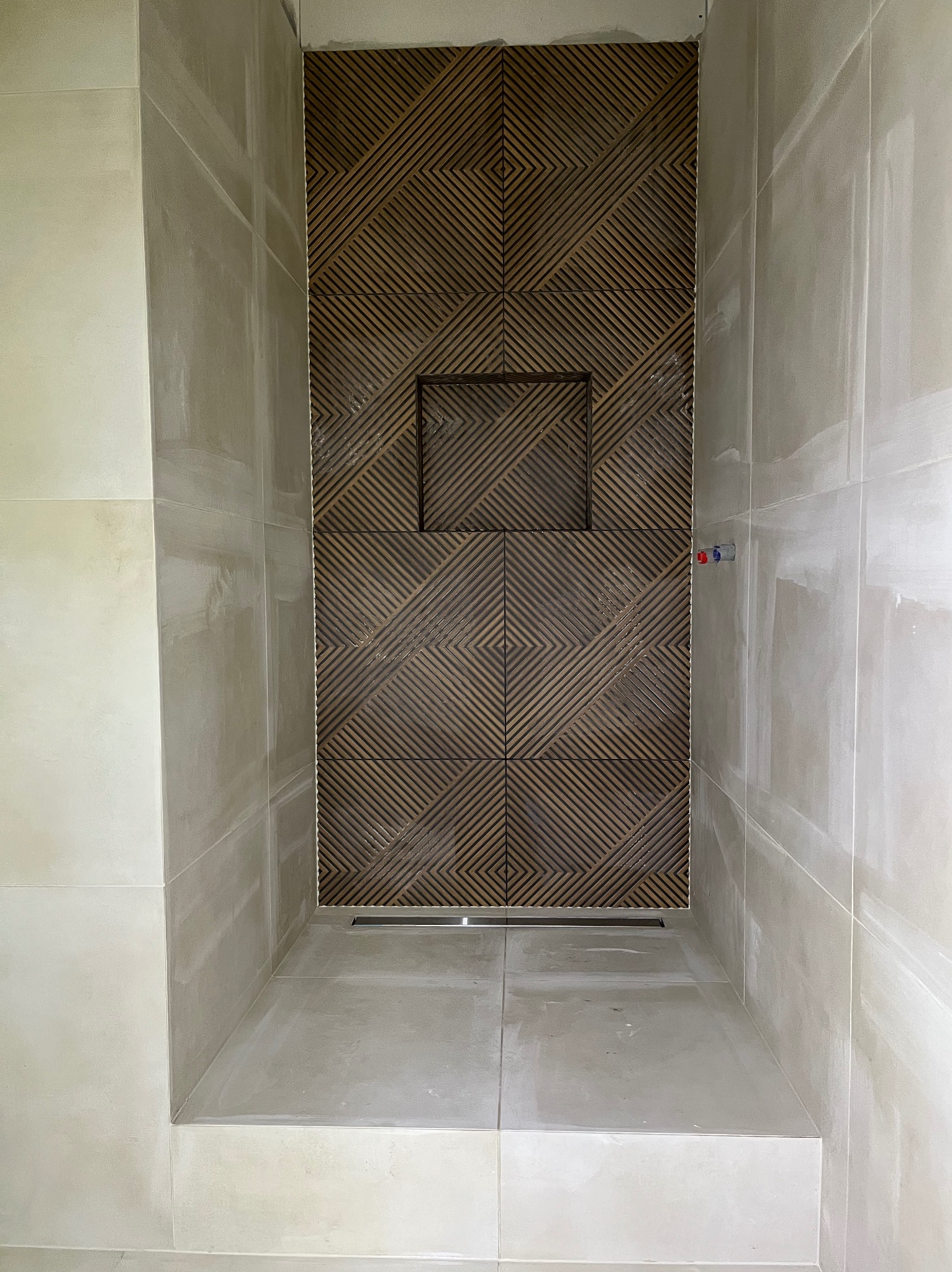 Wykończona kabina prysznicowa z dekoracyjną, drewnopodobną mozaiką i odpływem liniowym w Krosno. Jasne płytki na ścianach i podłodze, widoczne przyłącza wodne.