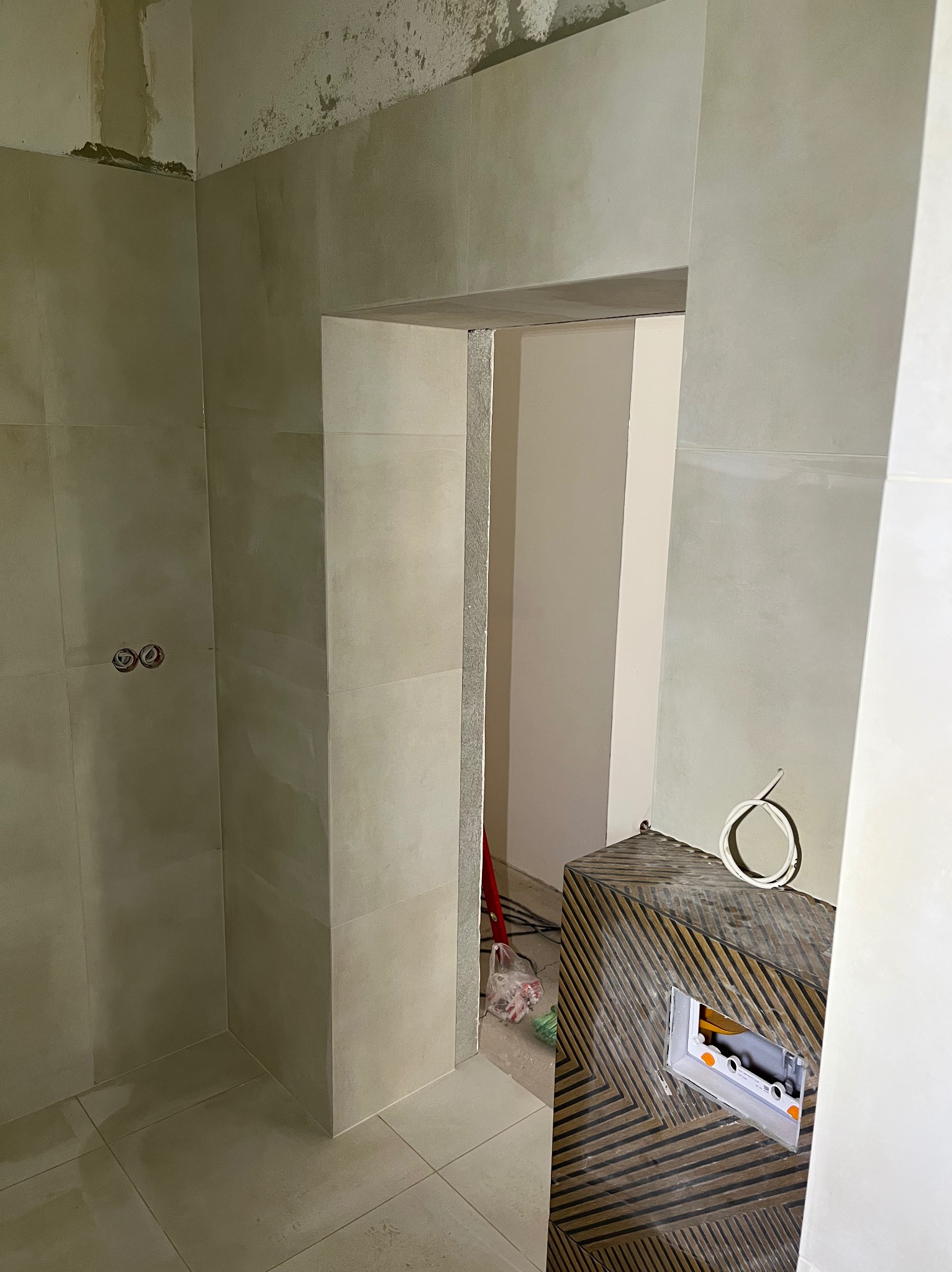 Wykończona łazienka z beżowymi płytkami, widoczne gniazdka elektryczne i fragment szafki z wzorem chevron. Prace w toku, sufit częściowo nie wykończony.