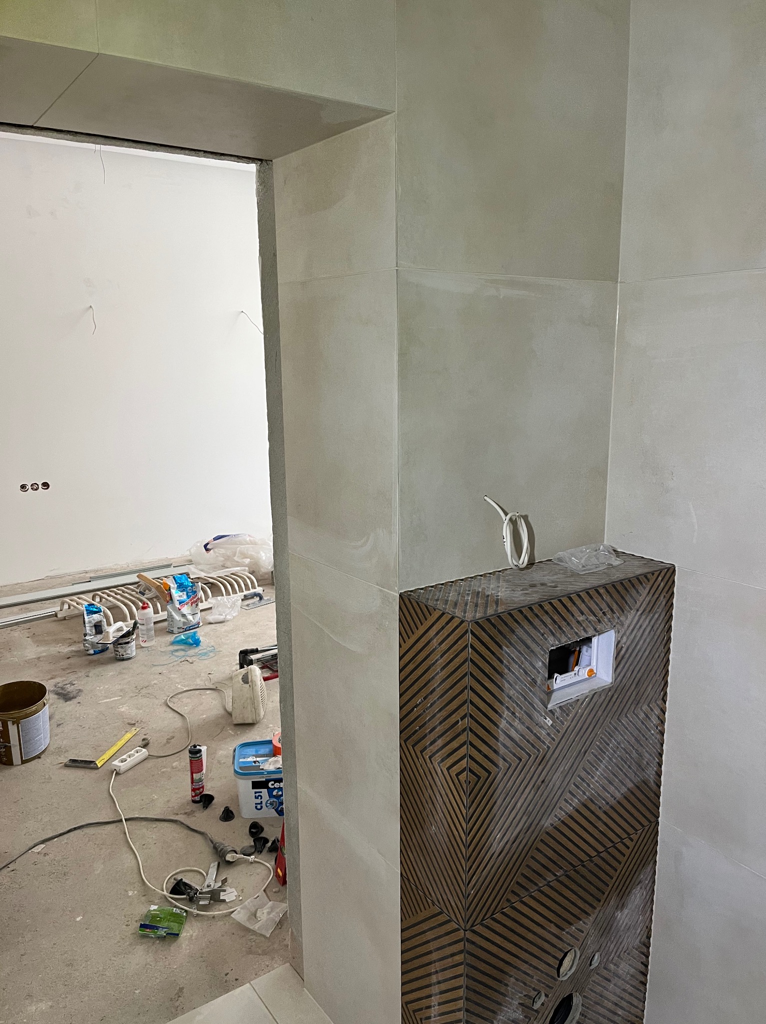 Wnętrze łazienki w trakcie wykończenia: obudowa stelaża WC z dekoracyjną okładziną, fragment ściany z jasnymi płytkami, widoczne narzędzia i materiały budowlane.