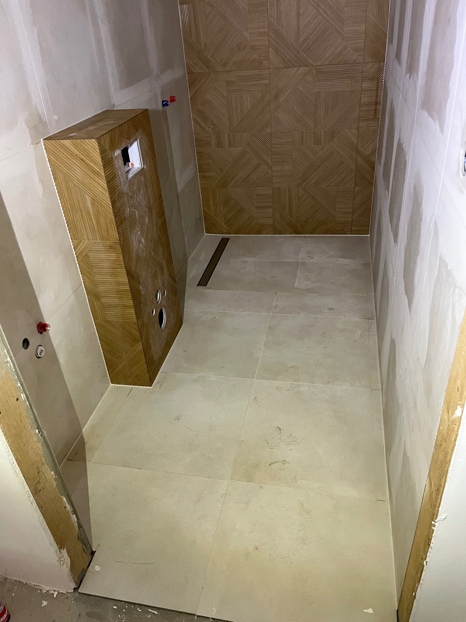 Wykończona łazienka z beżowymi płytkami na podłodze i ścianie prysznicowej. Zabudowa WC z drewnopodobną okładziną. Widoczne fragmenty ścian przygotowane do malowania.