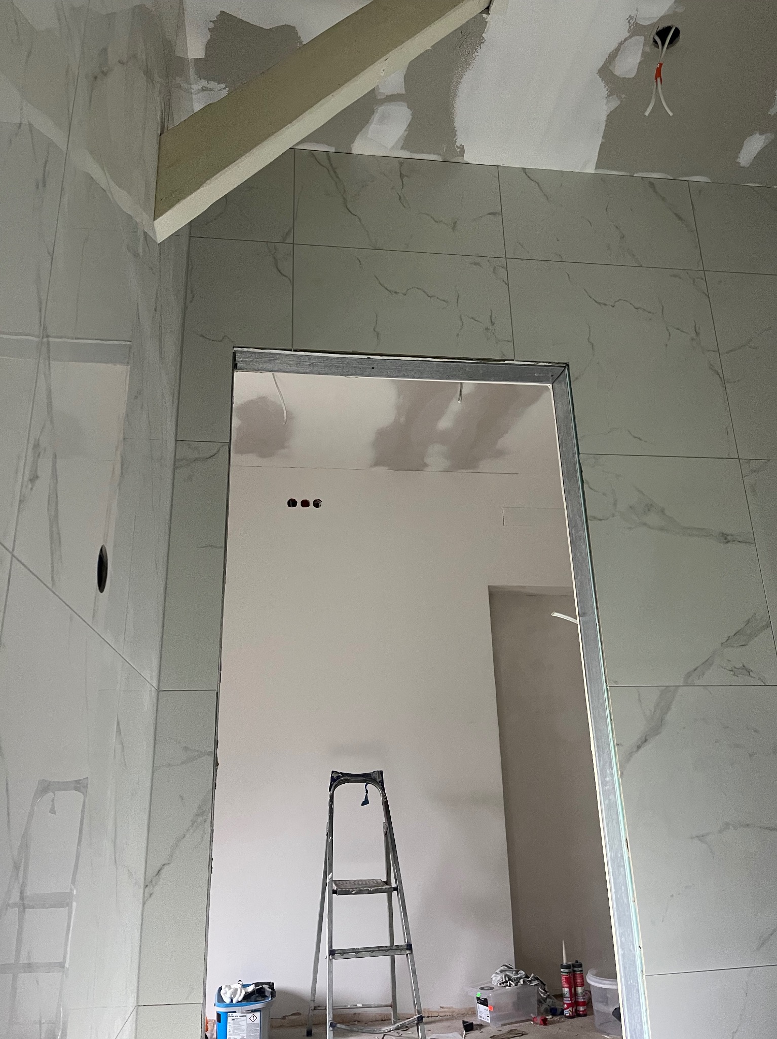 Łazienka w trakcie wykończenia: białe płytki imitujące marmur na ścianach, widoczna drabina, surowy otwór drzwiowy i częściowo wykończony sufit z instalacją.