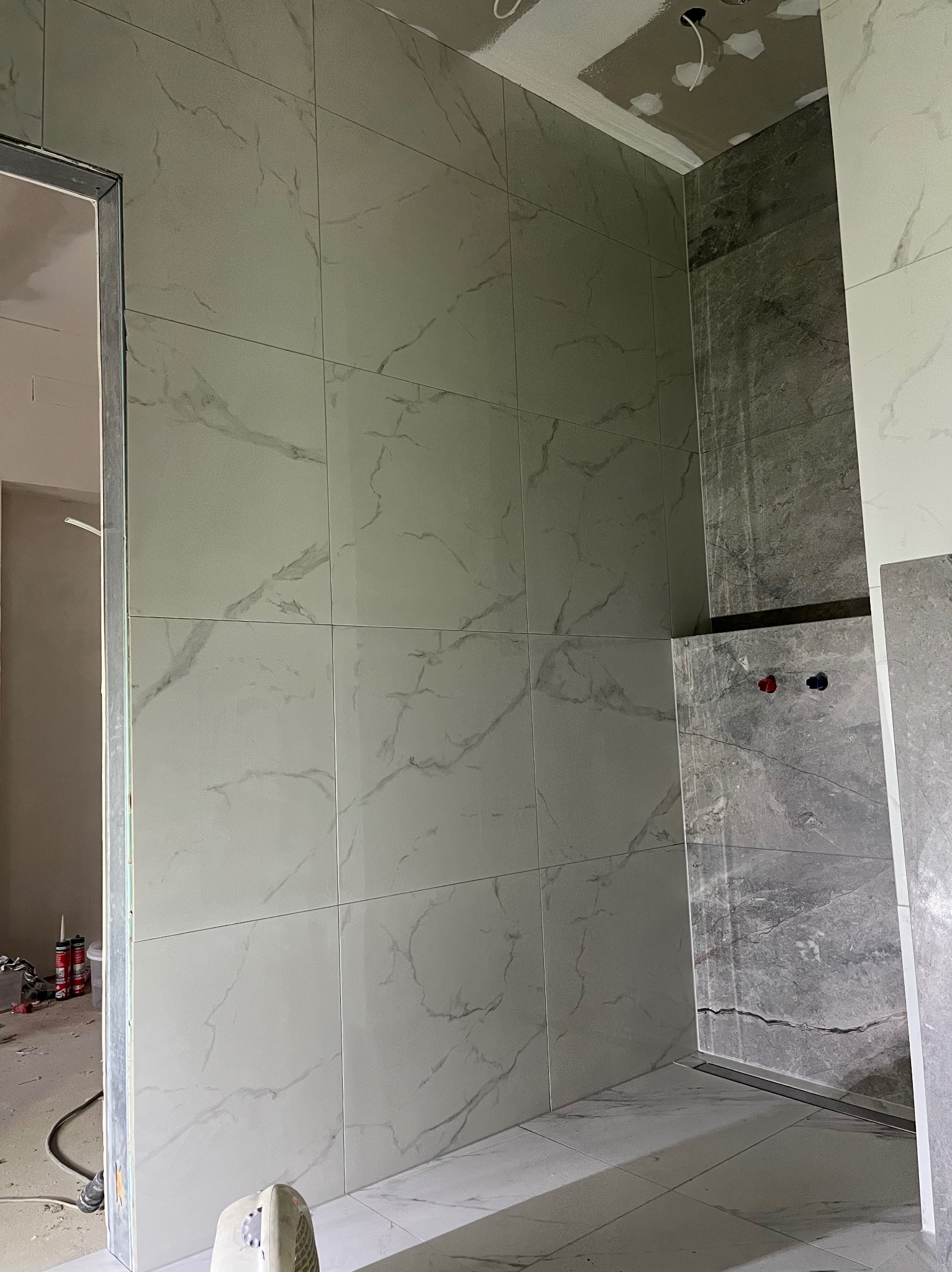 Ściany łazienki wyłożone jasnymi płytkami imitującymi marmur, z widocznymi przyłączami wodnymi i odpływem liniowym. Częściowo wykończona sufitowa płyta karton-gips.