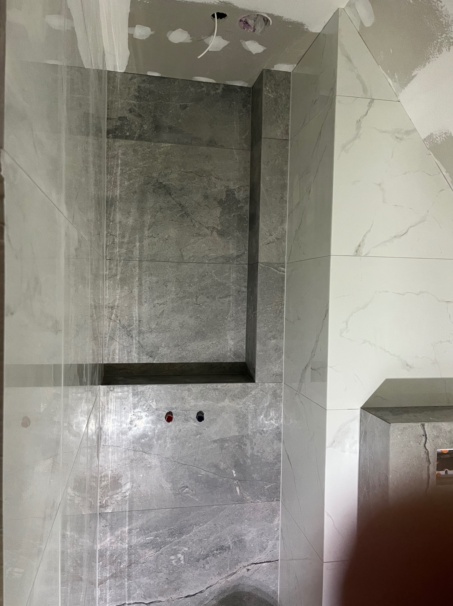 Wykończona wnęka prysznicowa z szarymi i białymi płytkami imitującymi marmur. Widoczne otwory na baterię i górna część kabiny. Ściany przygotowane do malowania.