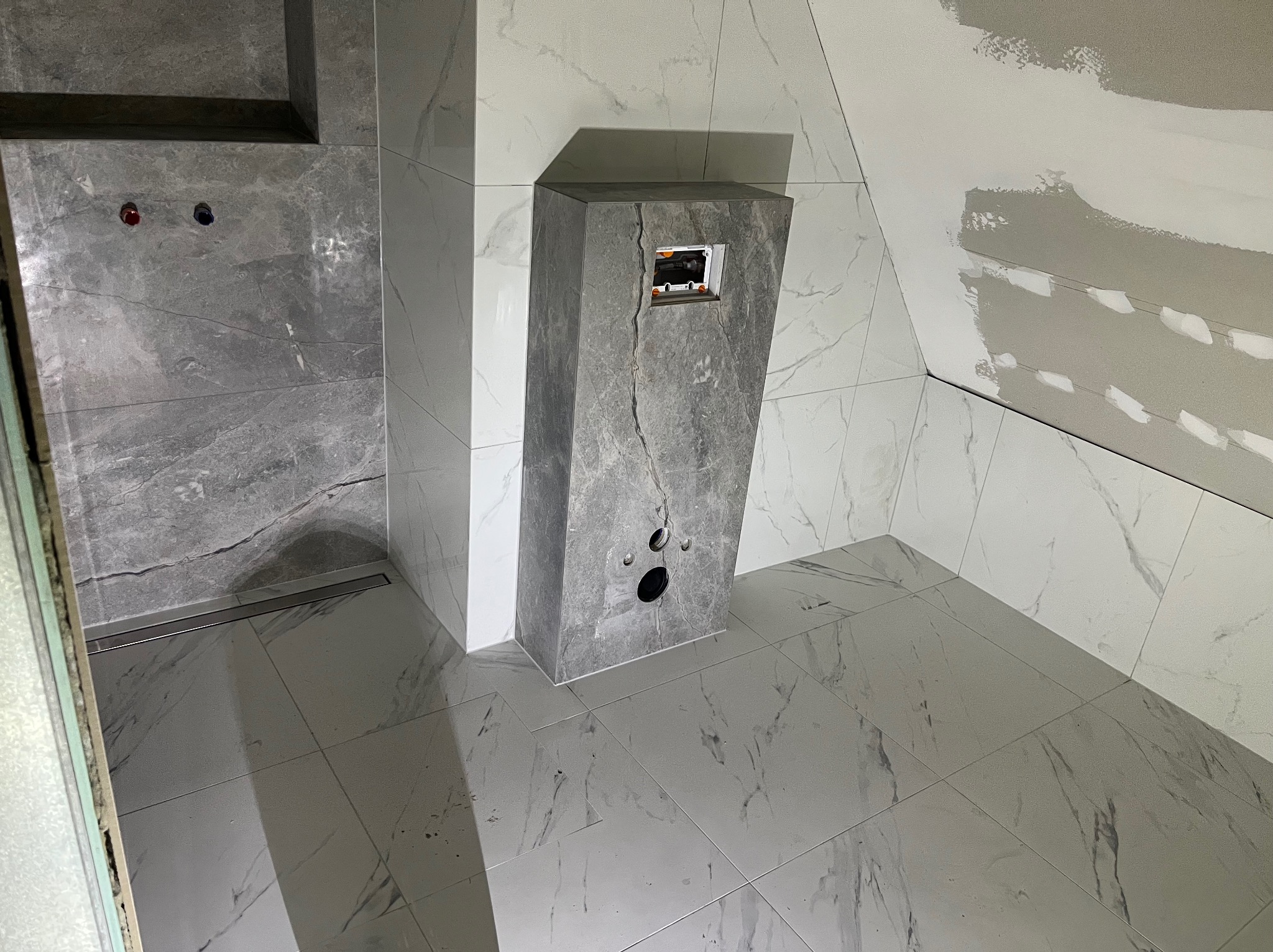 Wykończona łazienka w szaro-białej kolorystyce z marmurkowym wzorem płytek. Widoczna zabudowa stelaża WC i fragment ściany ze śladami szpachlowania.