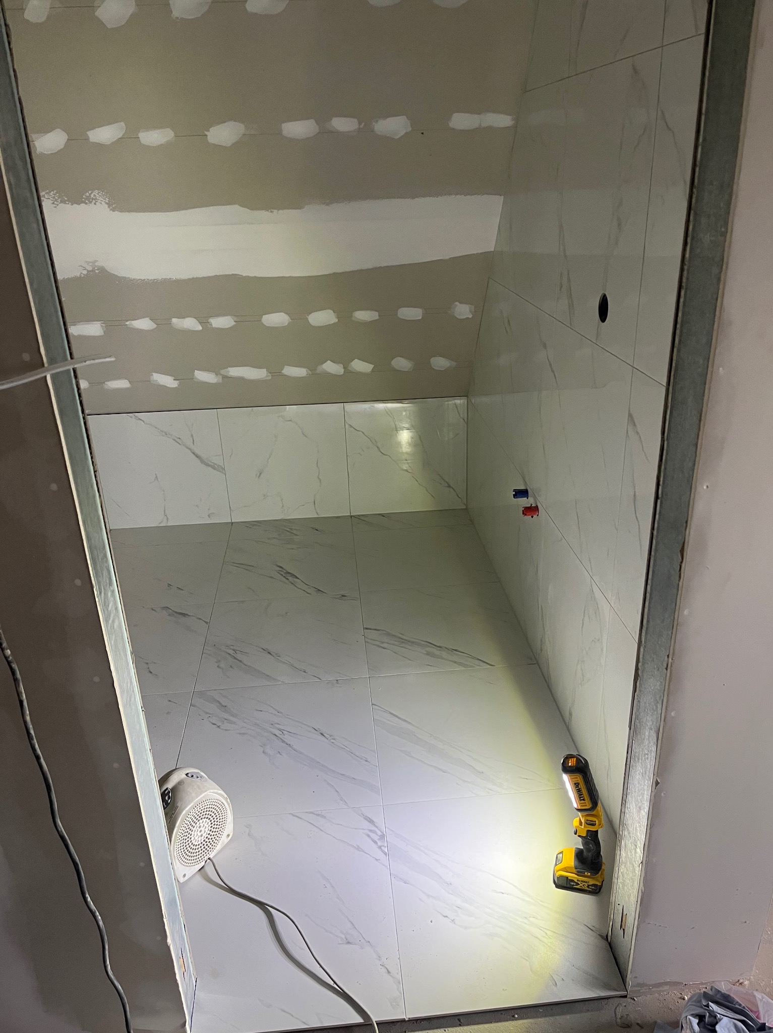 Wnętrze łazienki w trakcie prac: białe płytki imitujące marmur na ścianach i podłodze, sufit z płyt karton-gips, widoczne narzędzia i lampa budowlana. Etap wykończeniowy.