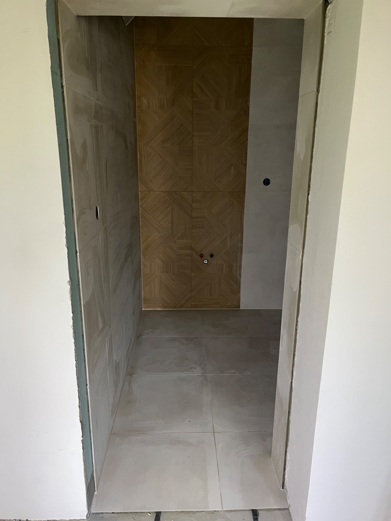 Wykończona łazienka z beżowymi płytkami o wzorze geometrycznym na ścianie i podłodze. Widoczne przyłącza wodne i otwór na gniazdko. Ościeżnica drzwiowa w trakcie montażu.