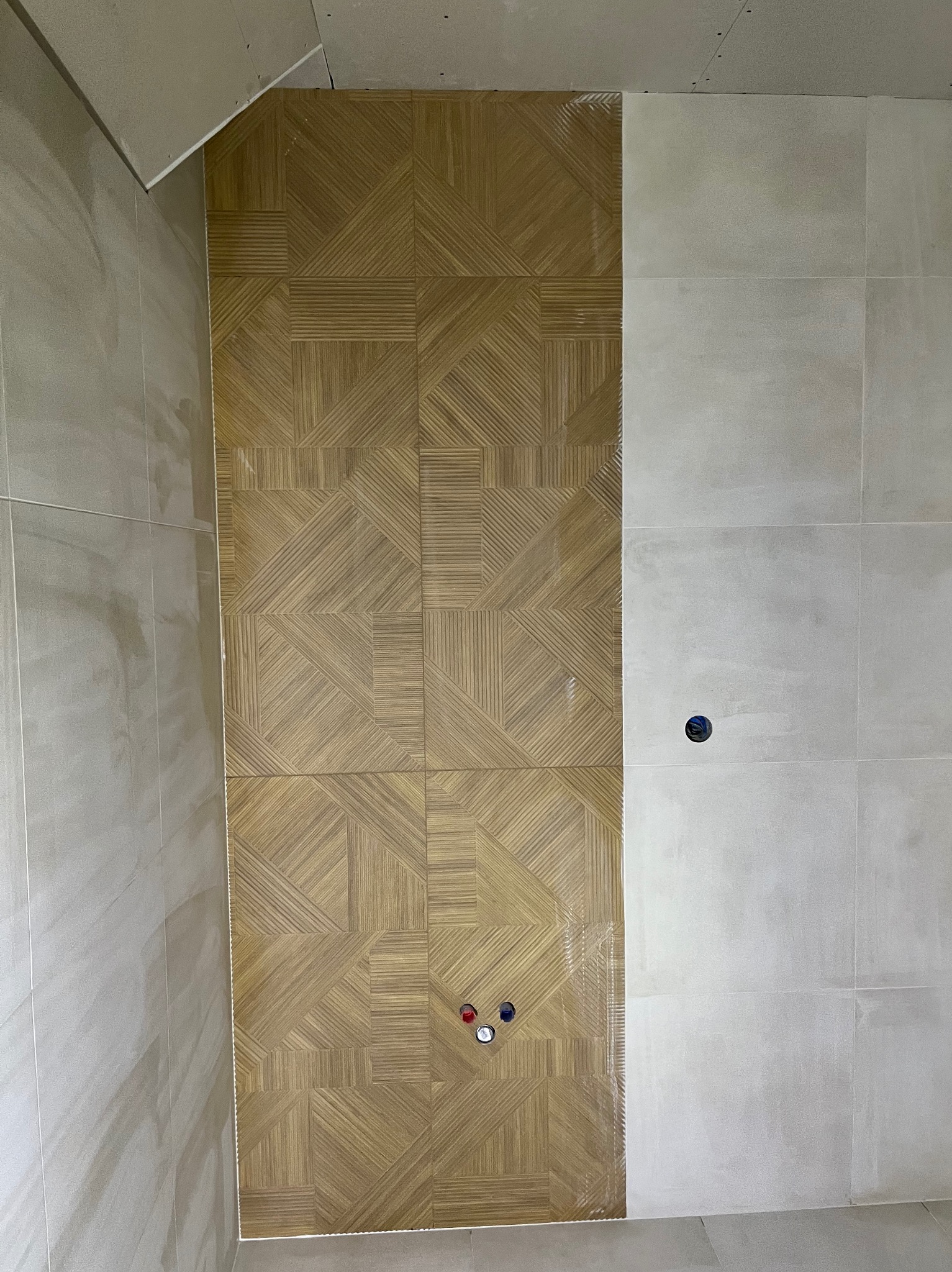 Ułożone płytki ścienne w łazience, z wzorem drewna po lewej i szare płytki po prawej. Widoczne przyłącza wodne i fragment sufitu z płyt kartonowo-gipsowych.