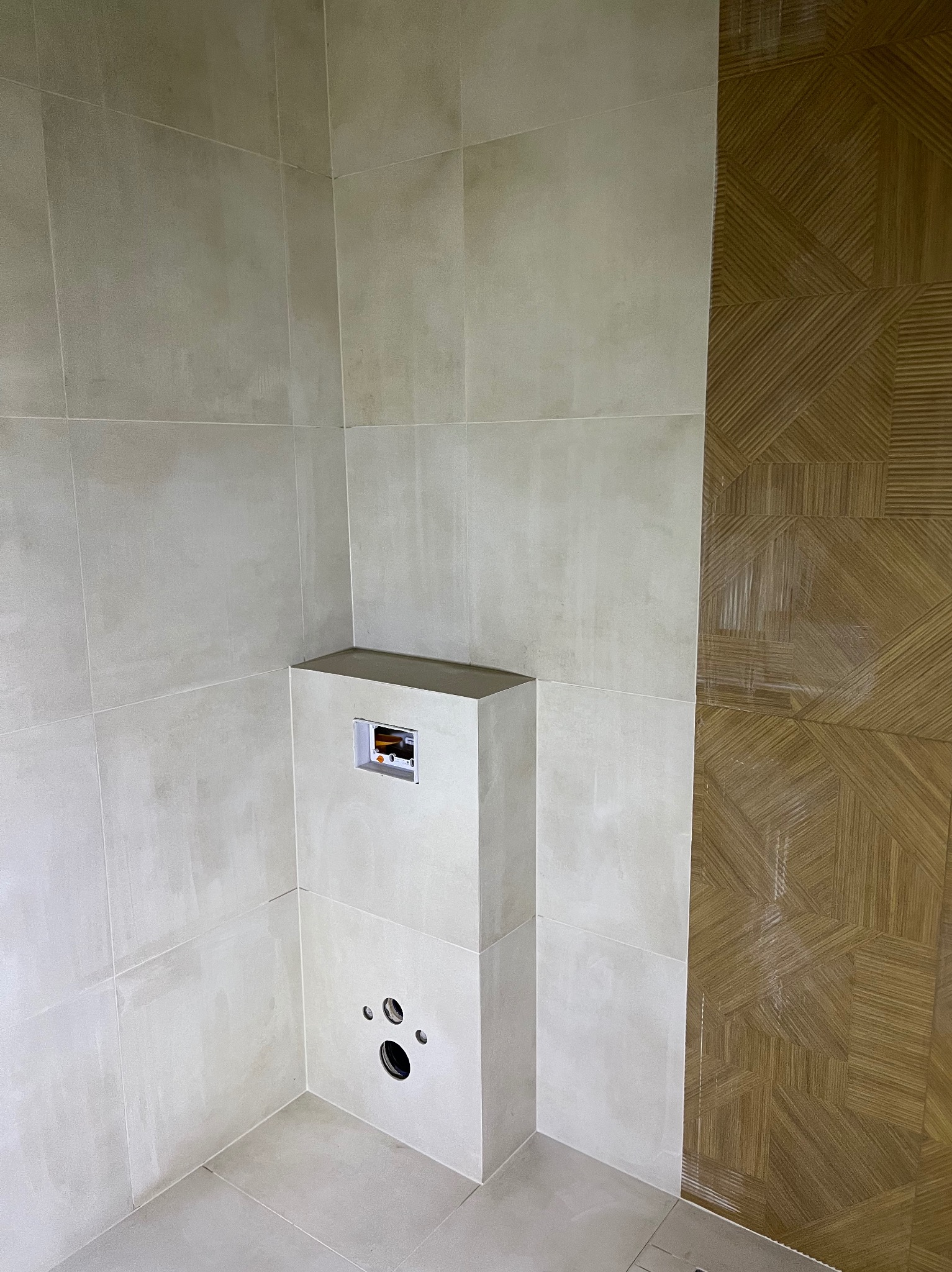 Wykończona łazienka z beżowymi płytkami i zabudową stelaża WC. Na ścianie dekoracyjne, brązowe płytki z geometrycznym wzorem. Widoczne otwory na przyłącza.