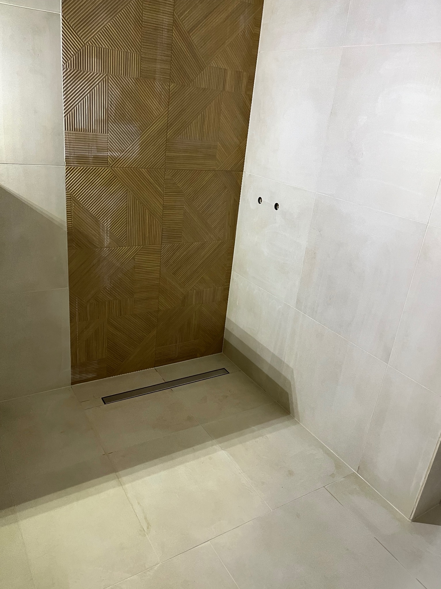 Wykończona strefa prysznicowa z odpływem liniowym i dekoracyjną, brązową ścianą z płytek o geometrycznym wzorze, kontrastującą z jasnymi płytkami na pozostałych ścianach.