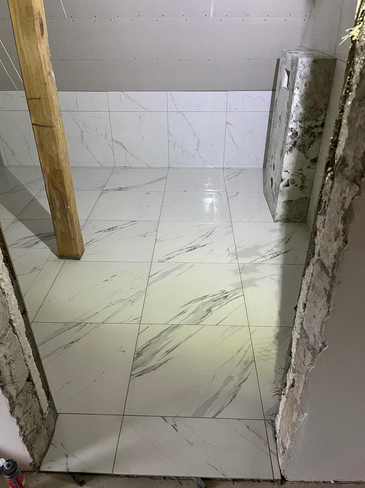 Ukończona podłoga i ściany w łazience wyłożone białymi płytkami imitującymi marmur, widoczne surowe wykończenie ścian i drewniana belka podtrzymująca sufit.