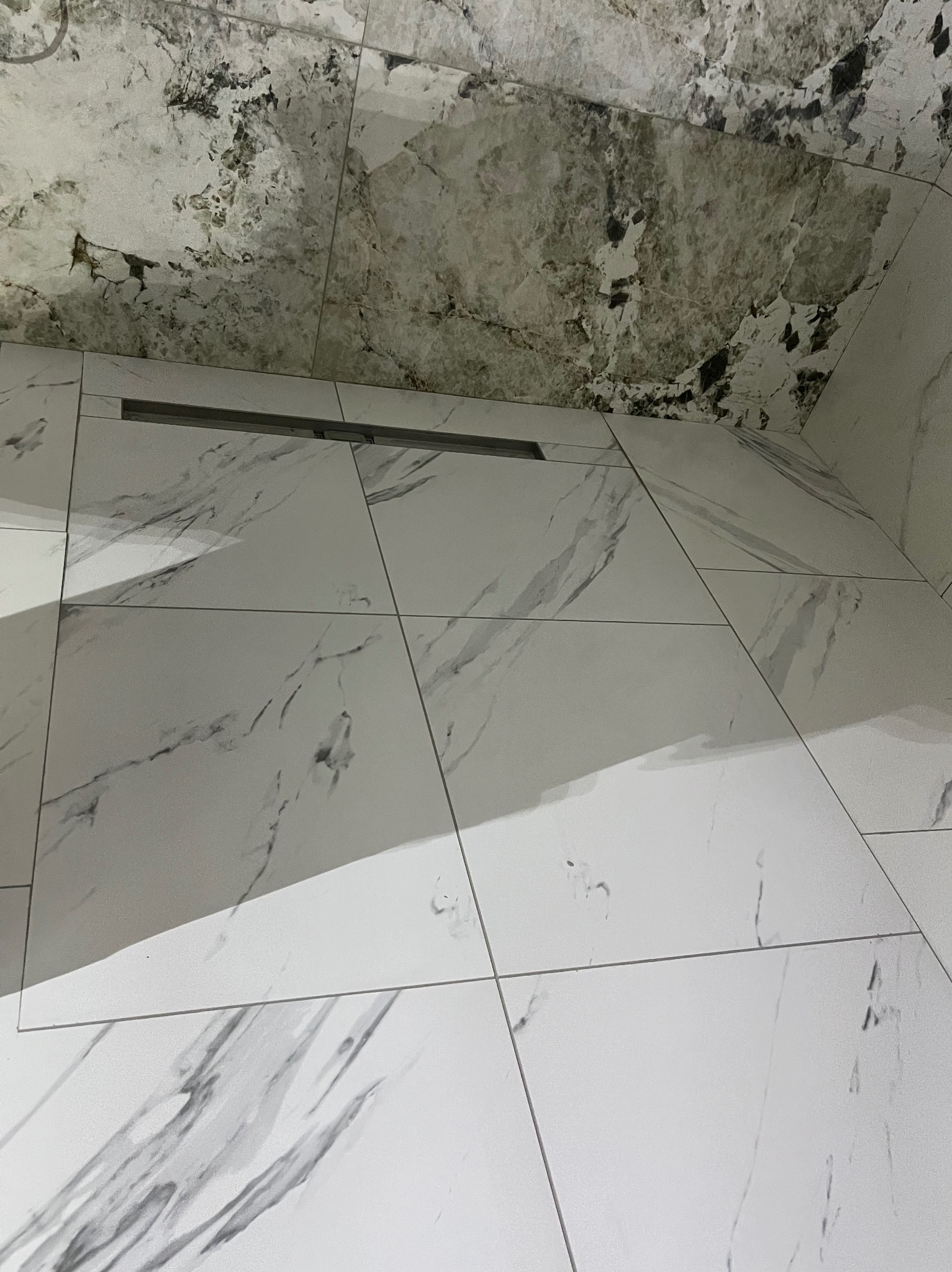 Białe płytki imitujące marmur na podłodze i ścianie łazienki, z widocznym odpływem liniowym. Górna część ściany z widocznymi śladami wilgoci i pleśni.